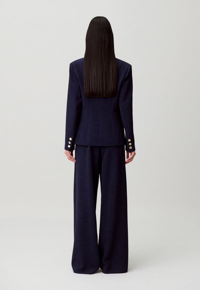 Completo sartoriale blu navy con blazer aderente decorato da bottoni dorati e pantaloni a gamba larga, presentato di spalle su uno sfondo neutro.