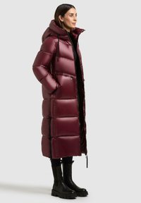 Lange bordeauxrode puffercoat met glanzende, gewatteerde textuur, een kap en ritszakken. Draagt over zwarte kleding met zwarte platformlaarzen.