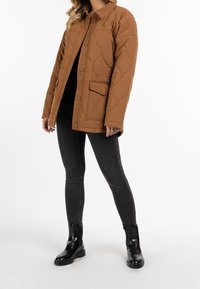 Veste matelassée marron avec fermeture à boutons sur le devant, manches longues et poche poitrine. Portée sur un haut noir et un jean gris avec des bottines noires.