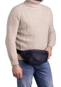Marsupio di tessuto di colore blu navy, con scomparto con zip, cinturino nero regolabile, indossato intorno alla vita. Maglione lavorato a maglia di colore crema e jeans di denim visibili.