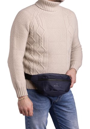 Fabric fanny pack in navy blue, zippered compartment, adjustable black strap, worn across waist. Knitted cream sweater and denim jeans visible.