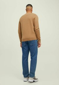 Vaaleanruskea neule pyöreällä kaula-aukolla, yhdistettynä sinisiin farkkuihin. Jalkineet ovat harmaat ja valkoiset tennarit. Näkymä on takaa.