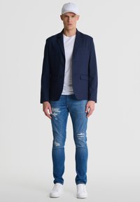 Jūros mėlynos spalvos bleiseris su dviem mygtukais, baltas marškinėlis, šlifuoti mėlyni džinsai ir balti sportbačiai, pasižymintys pritaikytu dizainu ir lygiu audinio paviršiumi.