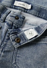 Vaqueros de mezclilla de un azul claro con un acabado texturizado. Cuentan con un botón metálico y una etiqueta de tela blanca con "Lil'Atelier" en la cinturilla.