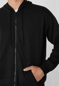 Felpa nera con zip realizzata in tessuto morbido, dotata di cappuccio con cordino, tasche anteriori e zip liscia con polsini a costine.