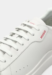 Zapatillas de cuero blancas con cordones blancos y un acabado texturizado. Presentan un logo rojo en el lateral y perforaciones sutiles en la puntera.