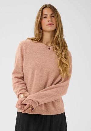 Femme aux longs cheveux blonds, portant un pull rose doux et une jupe noire, posant les mains jointes devant un fond uni.