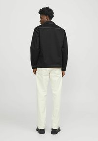 Black jacket with visible stitches, long sleeves, and a collar, paired with off-white pants and black shoes. Plain background.