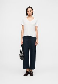 Γυναίκα που στέκεται φορώντας λευκό T-shirt με V-λαιμόκοψη, σκούρα φαρδιά τζιν, μαύρα mule με τακούνι και κρατώντας μια μαύρη τσάντα στο πλάι της.