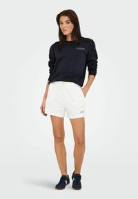 Mørkeblå sweatshirt og hvide shorts med elastisk talje. Shortsene har et logodetalje, sat sammen med mørke sneakers. Enkel, afslappet design.