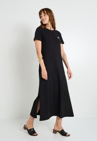 Robe maxi noire à manches courtes, fentes latérales et un petit détail de soleil brodé sur la poitrine. Le modèle porte des sandales noires à glissière.
