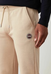 Jogging beige realizzati in tessuto morbido, dotati di vita regolabile con cordini e un patch circolare con il logo Colmar sulla coscia.
