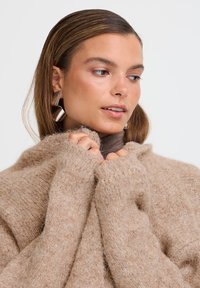 Maglione beige in lana con una finitura morbida e testurizzata, dotato di un collo alto e maniche ampie che coprono parzialmente le mani. Orecchini geometrici in argento.