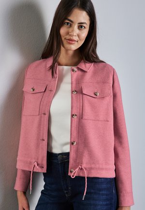Giacca di lana rosa con colletto, chiusura a bottoni, due tasche sul petto e orlo con cordino. Indossata sopra una maglietta bianca e abbinata a jeans scuri.