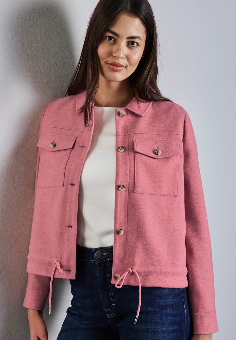 Veste en laine rose avec col, fermeture à boutons, deux poches sur la poitrine et ourlet à cordon. Portée par-dessus un haut blanc et associée à un jean foncé.