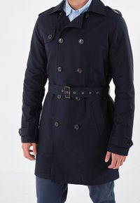 Marinblå trench coat i en klassisk dubbelknäppt design med bältesmidja, stora knappar och sidofickor.