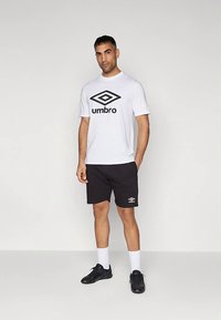 Bílé bavlněné tričko s černým logem "Umbro", doplněné černými sportovními šortkami s malým bílým logem, černými botami a bílými ponožkami.