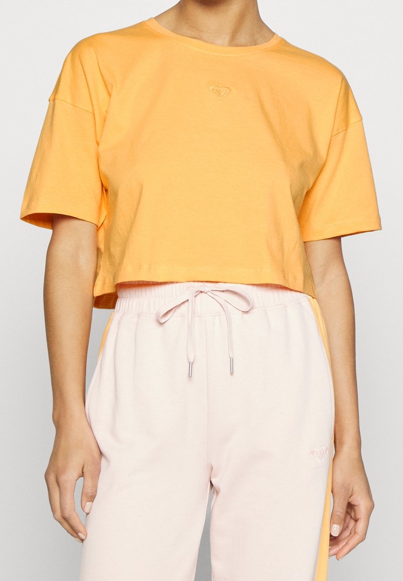 Osoba oblečená v volném oranžovém zkráceném tričku a světle růžových teplácích se stahovací šňůrkou, s oranžovými postranními pruhy a malými logy srdce.