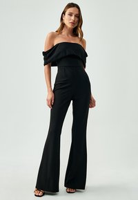 Sort off-shoulder jumpsuit med en tætsiddende overdel, brede flared ben og blød tekstur. Accentueret af en draperet halsudskæring og minimalistisk design.