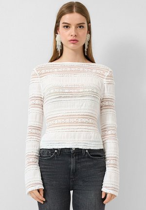 Woman wearing long-sleeve white lace top with horizontal patterns and black high-waist jeans, posing against plain light background.