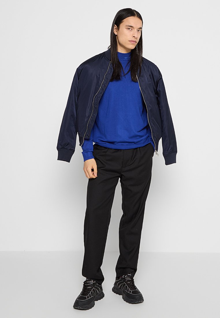 Marineblauer Bomberjacke über einem blauen Sweatshirt, kombiniert mit schwarzen Hosen und schwarzen strukturierten Sneakern. Einfaches Design mit elastischen Bündchen.