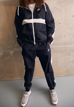 Osoba ubrana w czarno-biały dres Nike i białe buty sportowe Nike stoi oparta o beżową ścianę z rękami w kieszeniach.