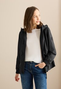 Svart lättviktsjacka med huva, dragkedja fram, elastiska muddar och två sidofickor. Bärs över en vit skjorta och blå jeans.
