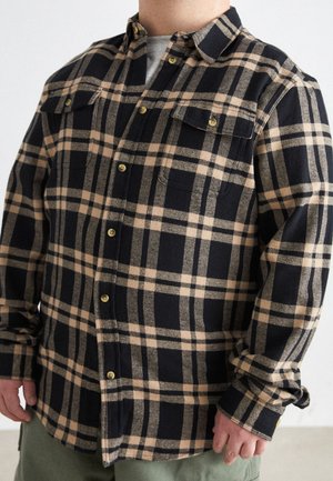 Mies, jolla on päällään ruskean ja mustan ruudullinen nappipaita harmaan t-paidan päällä sekä oliivinvihreät housut, seisoo käsien rentona sivuilla.