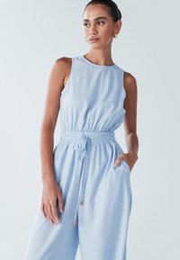 Svetlo modra brezrokavnik kombinezon z nabranim pasom, elastičnim trakom za zavezovanje in stranskimi žepi. Gladek material in sproščen kroj. Zlati kovinski detajli.