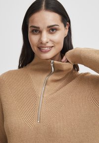 Pull zippé beige avec un col montant et un design texturé à côtes, doté d'une fermeture éclair argentée et d'une silhouette ajustée.
