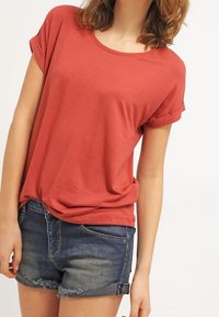 T-shirt couleur rouille avec un col rond et des manches courtes retroussées, associé à un short en denim déchiré. Tissu doux et léger.