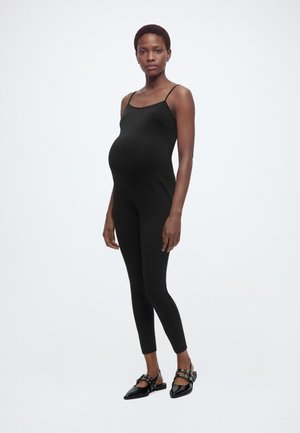 Schwangere Frau steht frontal, trägt einen eng anliegenden schwarzen ärmellosen Overall und schwarze spitze Schuhe mit Schnallen.