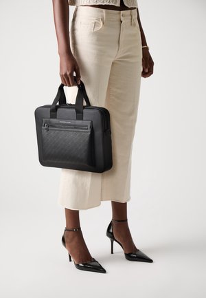 Persoon die crèmekleurige broek en zwarte hakken draagt en een zwarte gestructureerde handtas met een ritsvak aan de voorkant naast zich houdt.