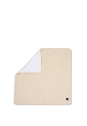 Couverture matelassée beige avec un motif géométrique subtil, un coin replié pour montrer le dessous blanc lisse, avec une petite étiquette de marque noire.