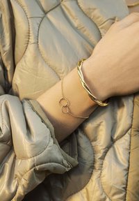 Bracciale rigido in oro attorcigliato e bracciale a cerchi intrecciati su un polso, posizionati su un morbido tessuto trapuntato grigio. Texture lisce con finiture lucide.