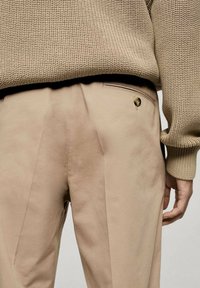 Beige broek van gladde stof, met plooien en een achterzak met een knoop. Draag het met een geweven, gebreide beige trui.