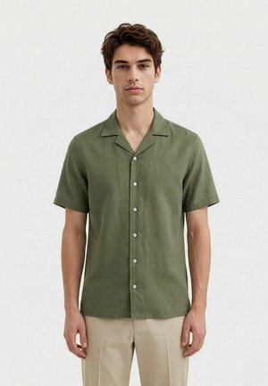 Ung mann som vender fremover, iført en olivengrønn kortermet knappeskjorte og beige bukser mot en ensfarget hvit bakgrunn.