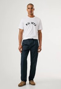 T-shirt branca de algodão com estampado preto "PJL 1973", combinada com calças azul-marinho. Ténis castanhos completam o conjunto. Design simples e casual.