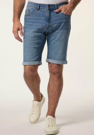 Mann trägt hellblaue hochgekrempelte Jeansshorts und weiße Sneaker, steht mit einer Hand in der Tasche und der anderen Hand entspannt.