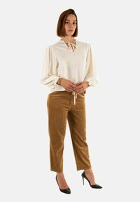 Blusa blanca a rayas con mangas abullonadas y un lazo en el cuello, combinada con pantalones de pana marrón y tacones altos negros.