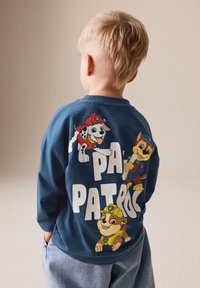Zila garām piedurknēm krekls ar košās krāsās attēlotiem karikatūru suņiem uz muguras. Uzraksts baltā krāsā "PAW PATROL" un mīksta tekstūra.