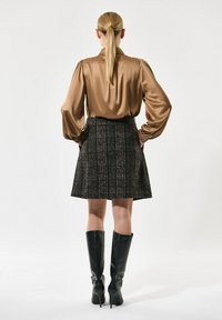 Blusa larga de satén marrón con mangas largas, combinada con una falda de tweed negra con un patrón de cuadros y botas de tacón altas negras hasta la rodilla.