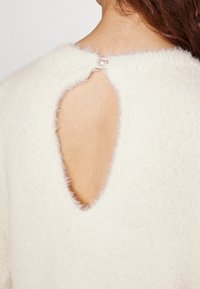 Vue arrière d'une personne portant un pull blanc duveteux avec une découpe en forme de goutte d'eau fermée par un petit bouton en perle au niveau du cou.