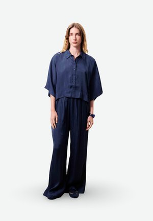 Femme debout, portant une chemise bleu marine ample à boutons avec des manches larges et un pantalon large assorti sur un fond uni.