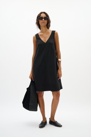 Woman wearing sleeveless black V-neck dress, black flats, sunglasses, and silver bracelet, holding a large black tote bag.