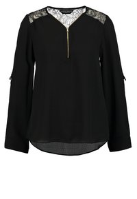 Blouse noire à manches longues avec panneaux en dentelle aux épaules et fermeture éclair dorée d’un quart de longueur sur l’encolure avant.