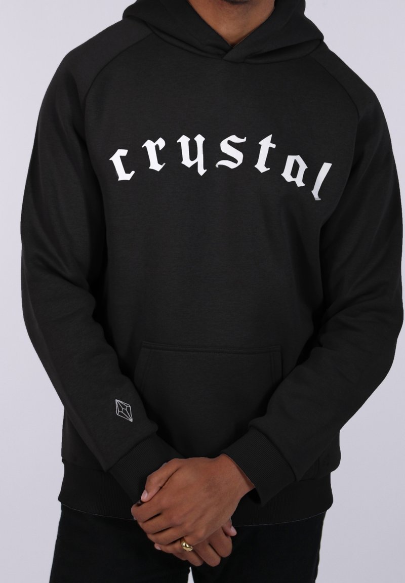 Personne portant un sweat à capuche noir avec un texte blanc en style gothique « crystal » sur la poitrine et un petit logo géométrique en forme de cristal sur la manche.