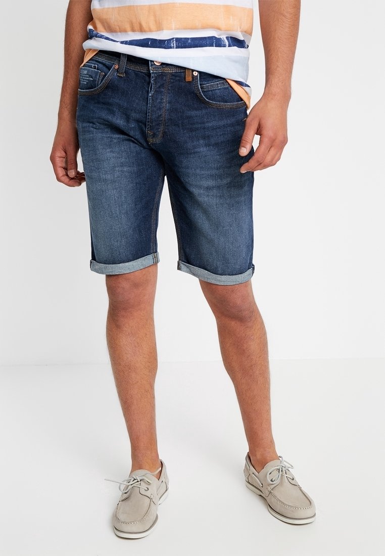 Džínové šortky v tmavém odstínu s ohrnutými lemy, doplněné pruhovaným tričkem v modré, oranžové a bílé barvě. Obuv tvoří světle šedé lodní boty.