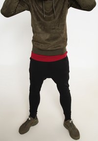 Felpa verde oliva con tasca frontale, indossata sopra una maglietta rossa, abbinata a pantaloni neri a sigaretta e sneakers verdi.