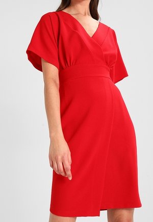 Femme portant une robe rouge vif longueur genou, avec des manches courtes larges et un corsage croisé en V.